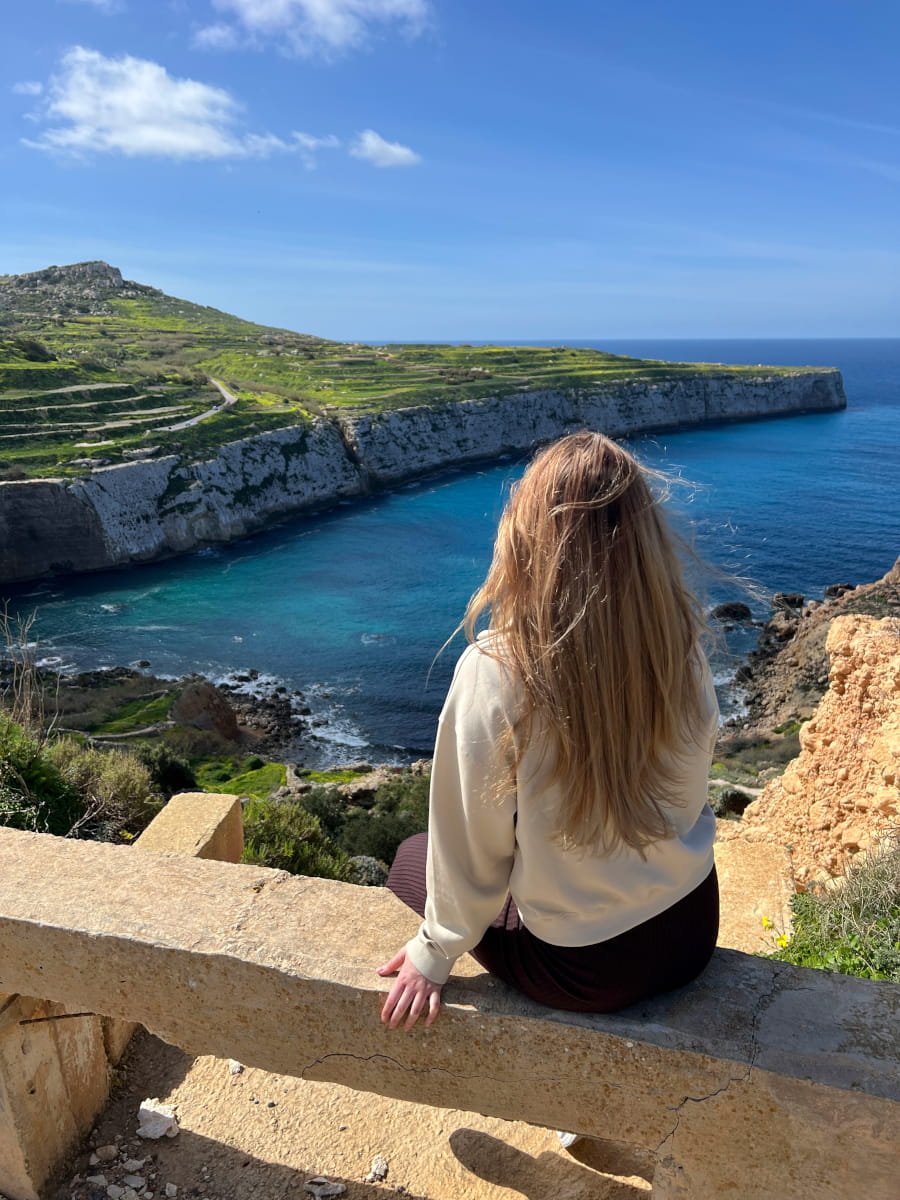 Sara Claeys die uitkijkt over een rotsachtige klif in Malta, als symbool voor het vinden van rust en een nieuw perspectief na de 'hustle'-cultuur.